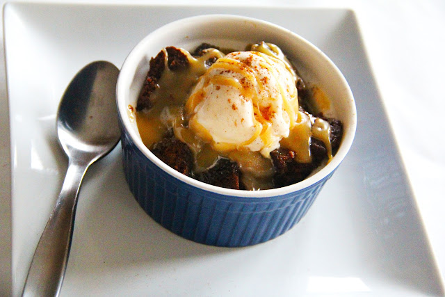 Sticky toffee gingerbread bread pudding topped with a scoop of vanilla ice cream in a small blue bowl with a silver spoon on the side.