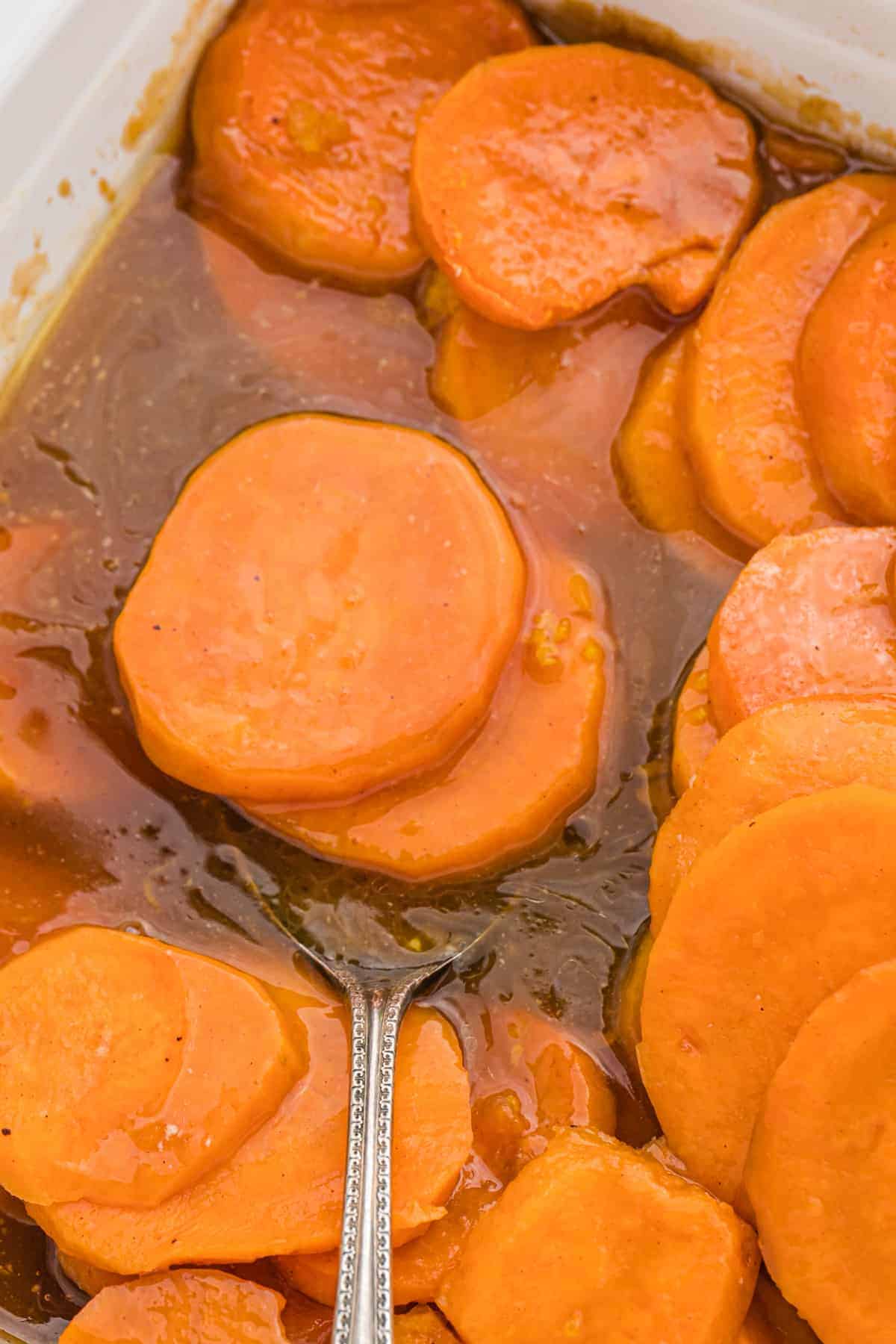A silver spoon in a serving dish of baked candied yams in brown sugar syrup