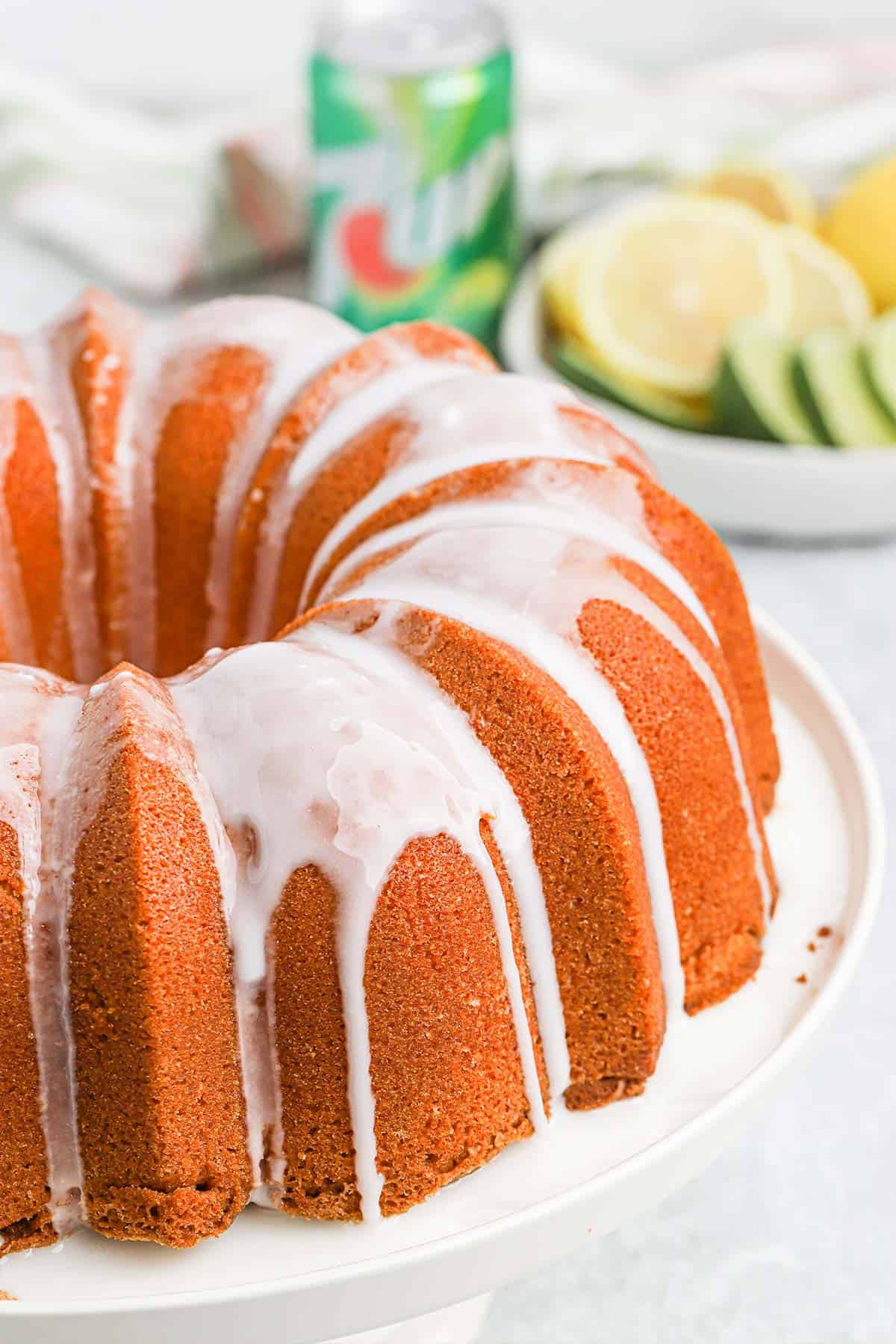 7up cake on a white pedestal with limes, lemons and a can of soda in the background.