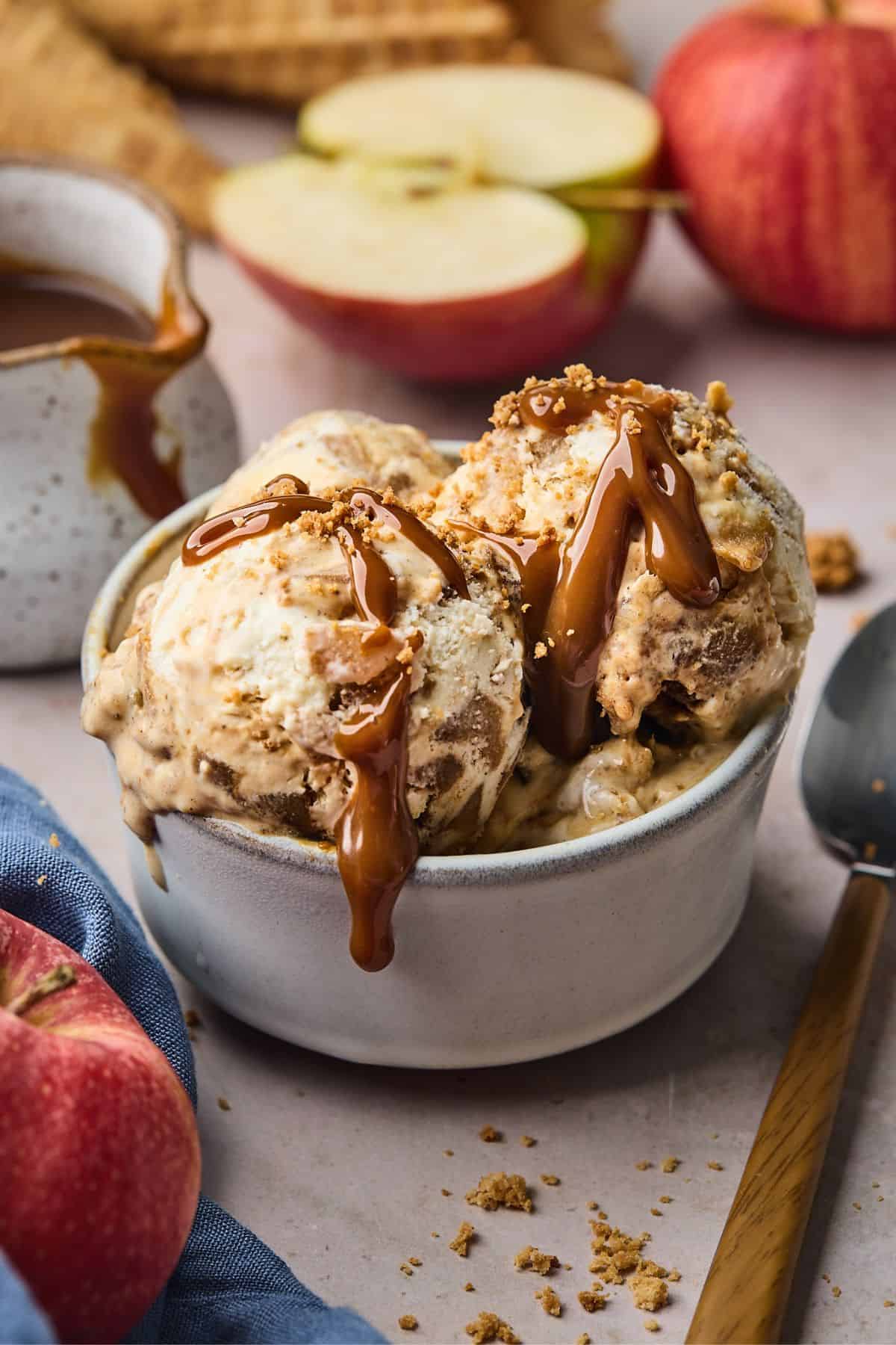 A bowl of apple pie ice cream with caramel sauce and red apples in the background