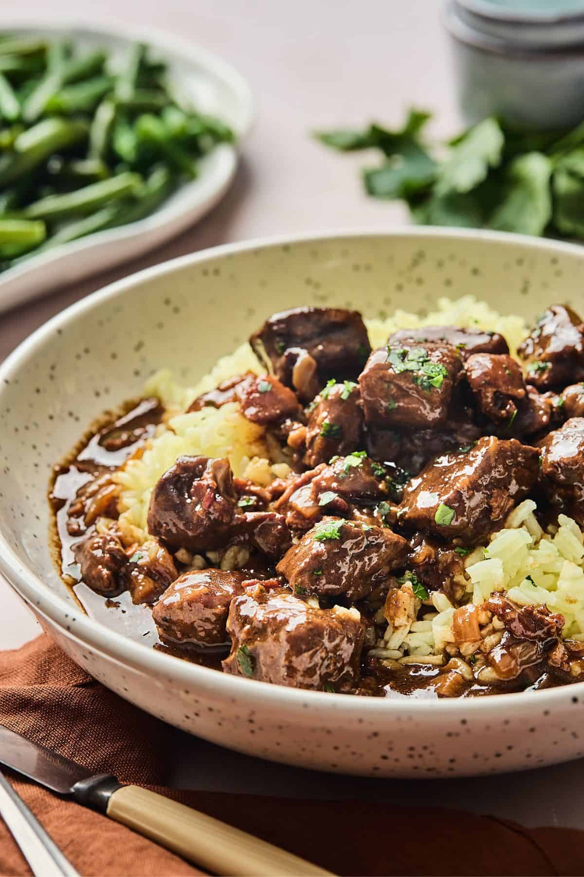 Beef tips over butter rice on a white plate with green beans in the background