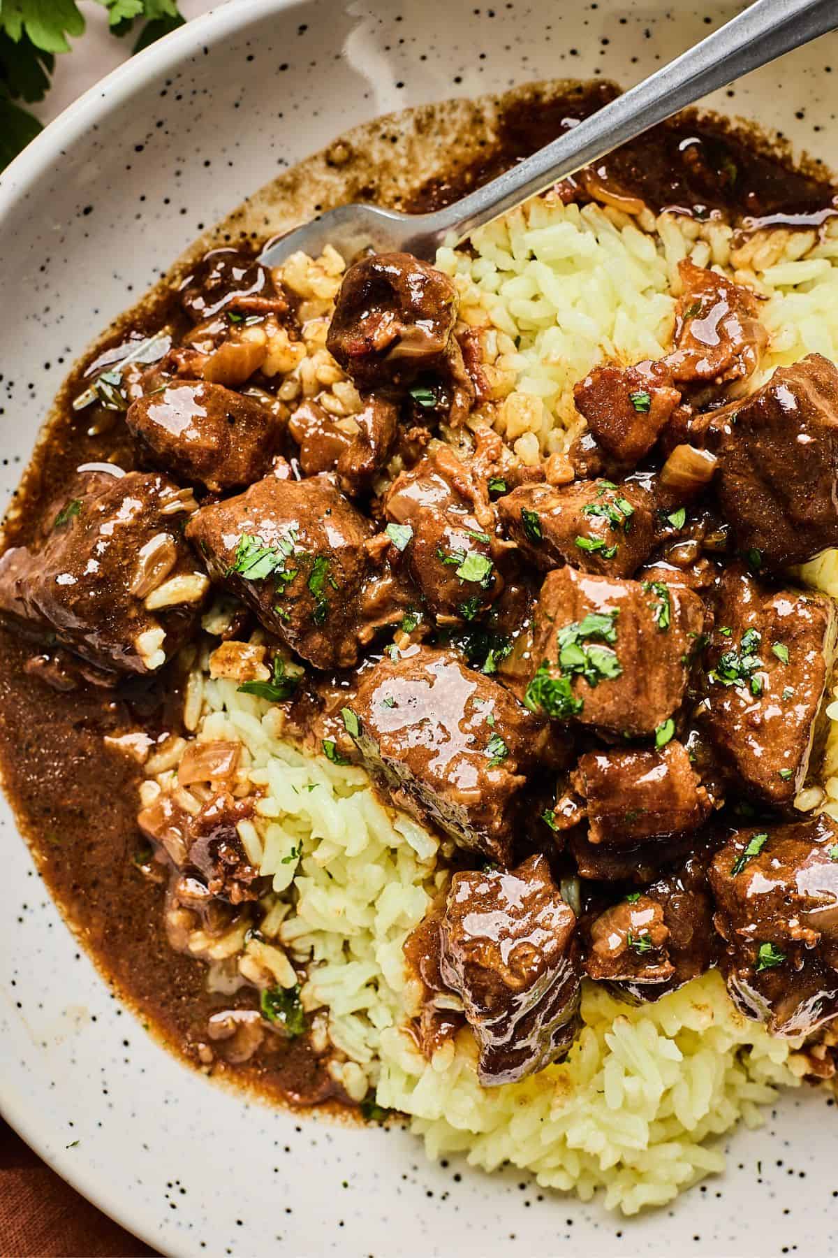 Beef tips topped with herbs over butter rice on a white plate with a fork