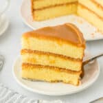 A slice of the best caramel cake recipe on a white plate under a cake server with full caramel cake in the white background