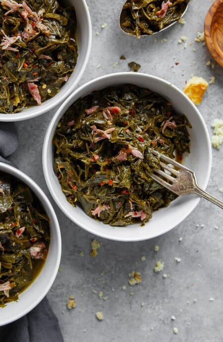 Overhead of three white bowls filled with Southern Collard Greens with hamhock against gray background