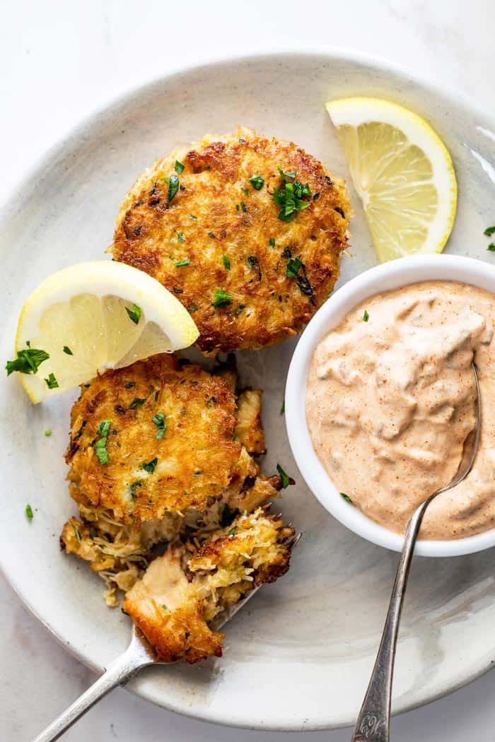 Seafood cakes being cut into to eat and served with lemon wedges and remoulade sauce