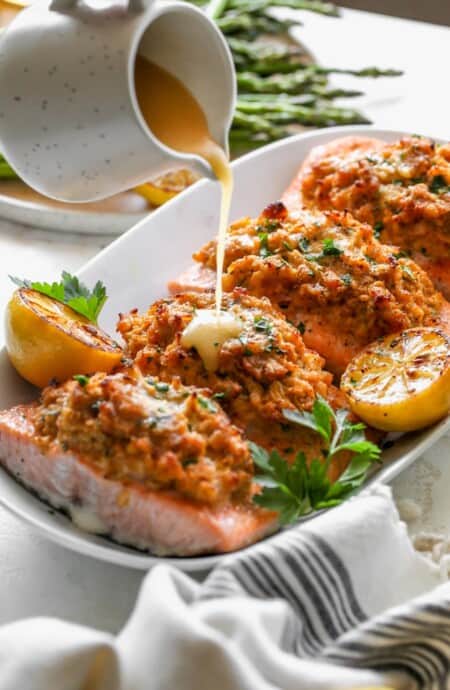 Crab stuffed fish recipe on a white platter with lemon sauce being poured on top in white background