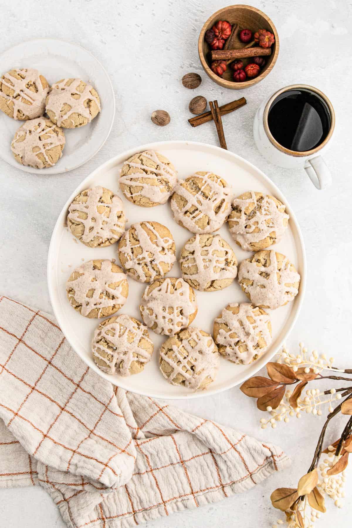 Eggnog Iced Butter Cookies A table top with a plate of eggnog butter cookies on top, spices, a tea towel, and cinnamon sticks.