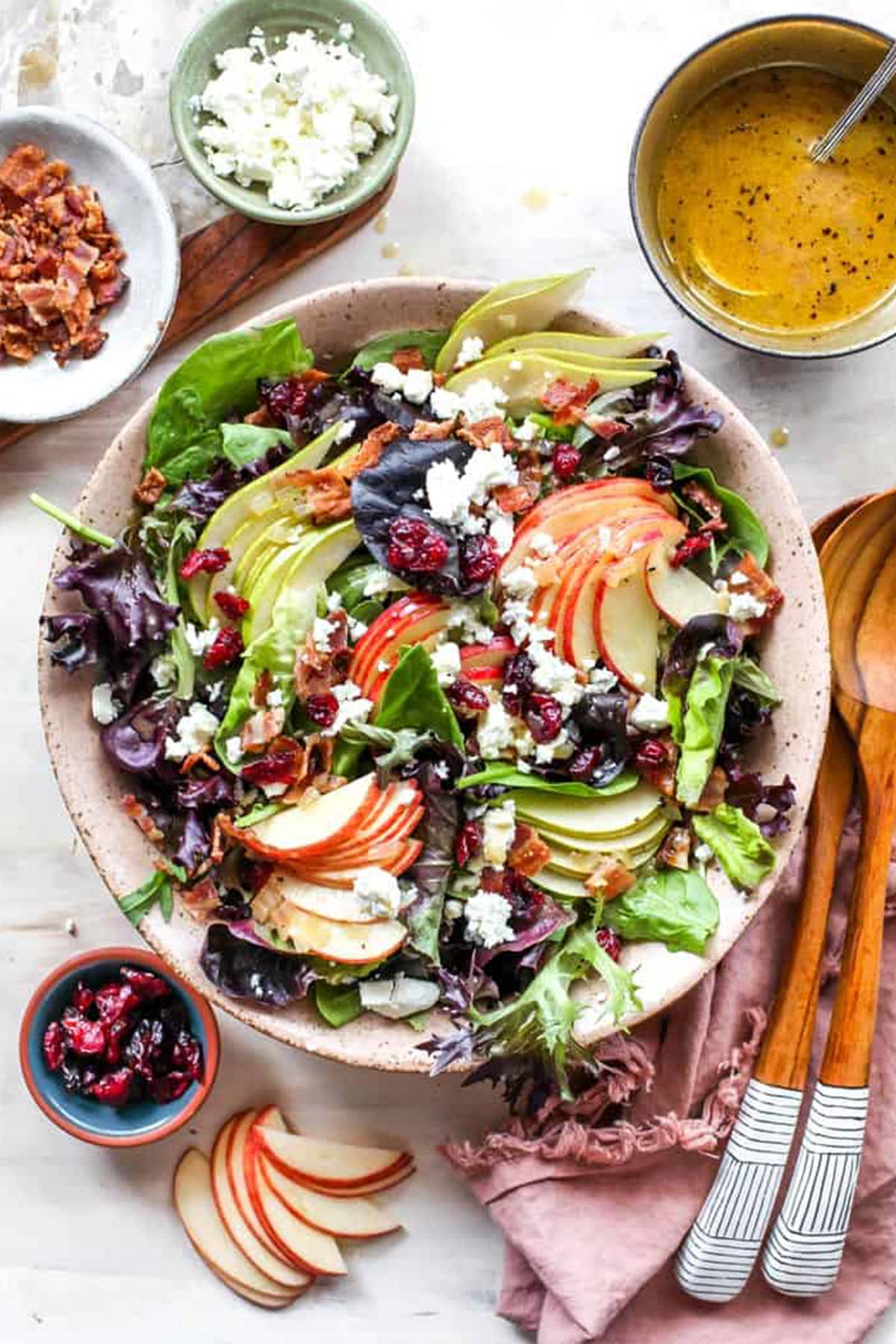A pink bowl of autumn salad surrounded by dried cranberries, apple cider vinegarette and bacon with spoons nearby