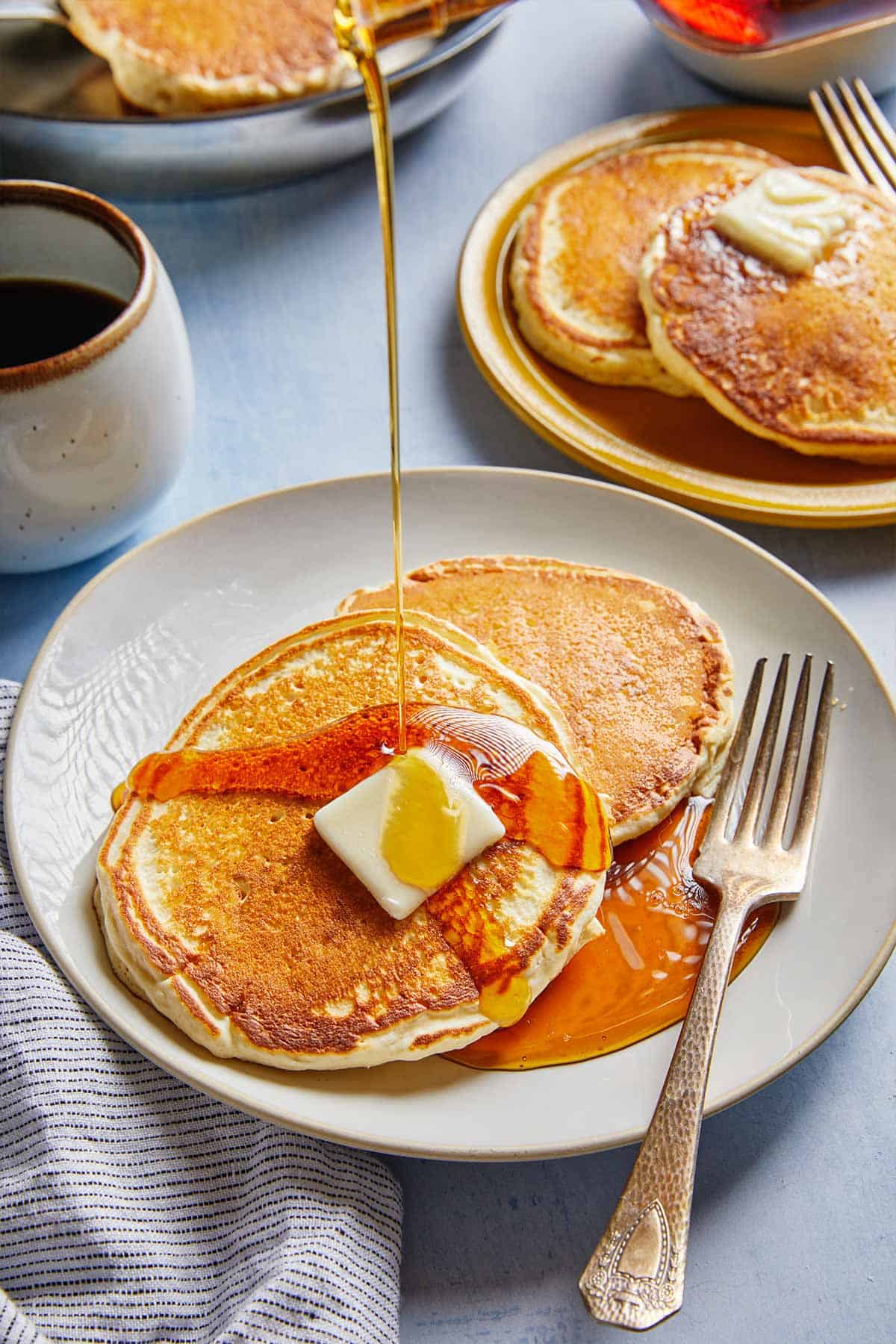 Pouring maple syrup from a pitcher over a plate of pancakes on the table.