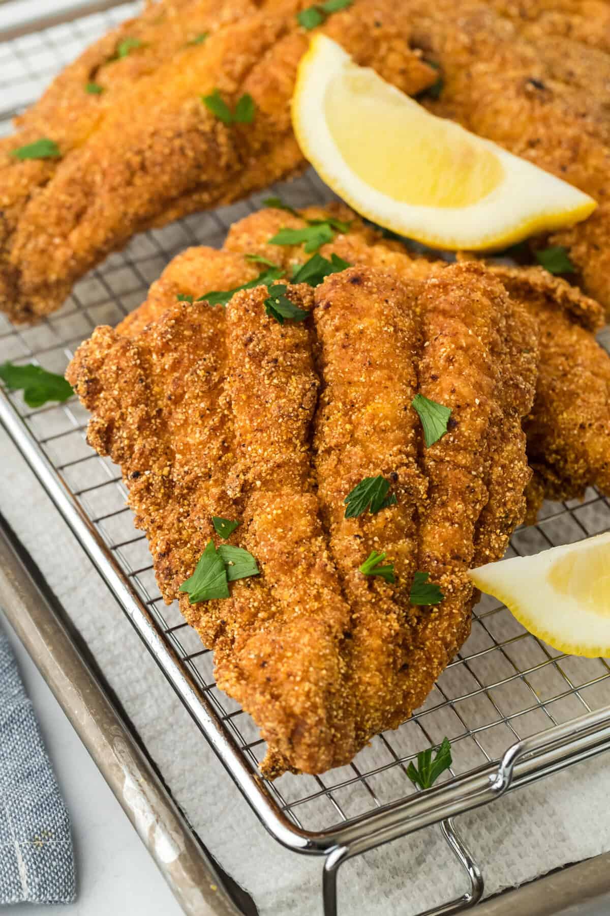 A close up of deep fried catfish on a wire rack with lemon wedges and pasley sprinkled on top