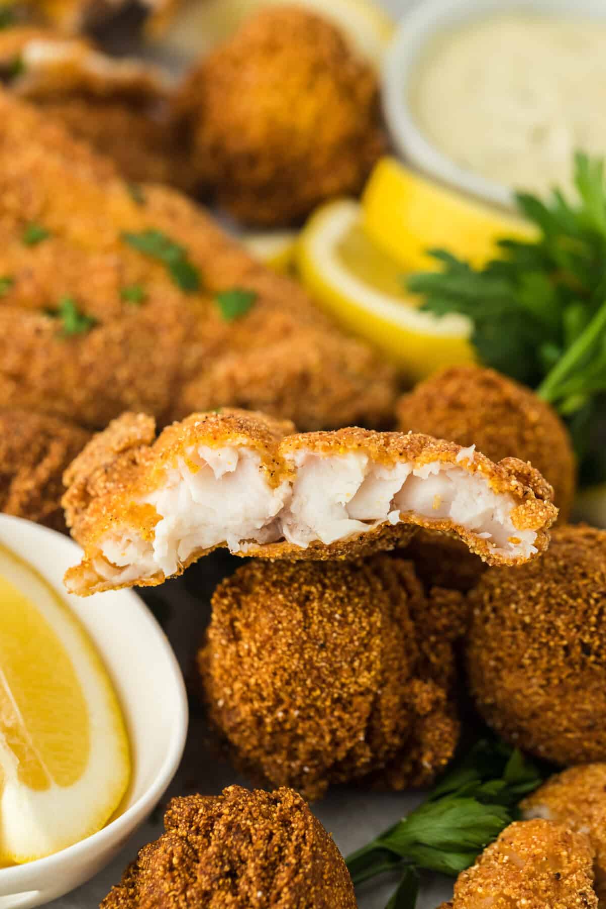 A close up of fried catfish being cut into so you can see the flaky inside with hush puppies and lemon in the background