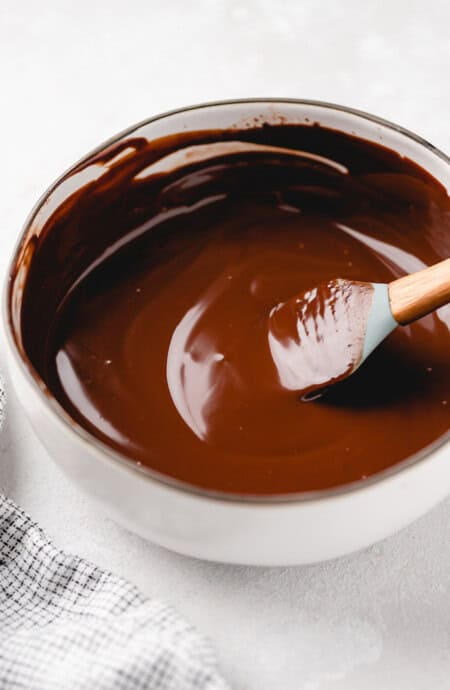A spatula stirring a smooth chocolate ganache in a white pot on a white background