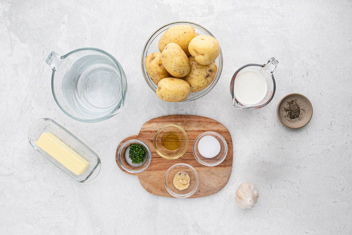 Ingredients in clear bowls like butter, yukon potatoes, seasonings, half and half on a white background to make easy garlic mashed potatoes recipe