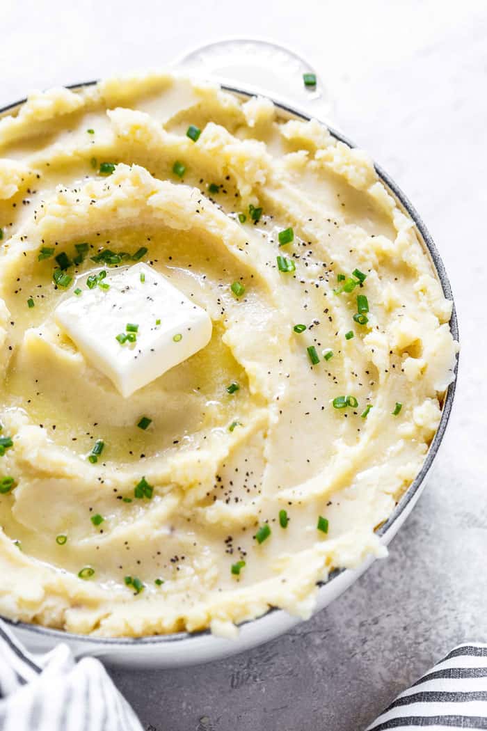 A close up of large bowl of roasted garlic mashed potatoes with pats of butter and parsley on a white gray background