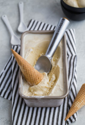 A large frozen container of homemade vanilla ice cream with waffle cones and a scooper ready to serve against striped napkin