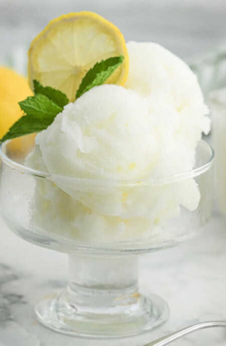 A glass dessert cup with two scoops of lemonade sorbet garnished with lemon slice and fresh mint.