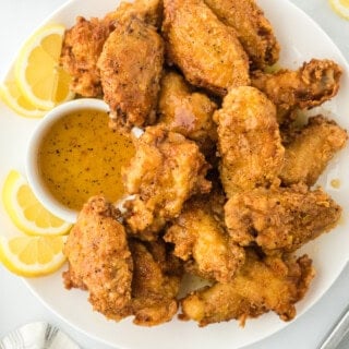 Fried lemon pepper wings on a white plate with lemon wedges and honey lemon pepper wet sauce in a bowl against white background