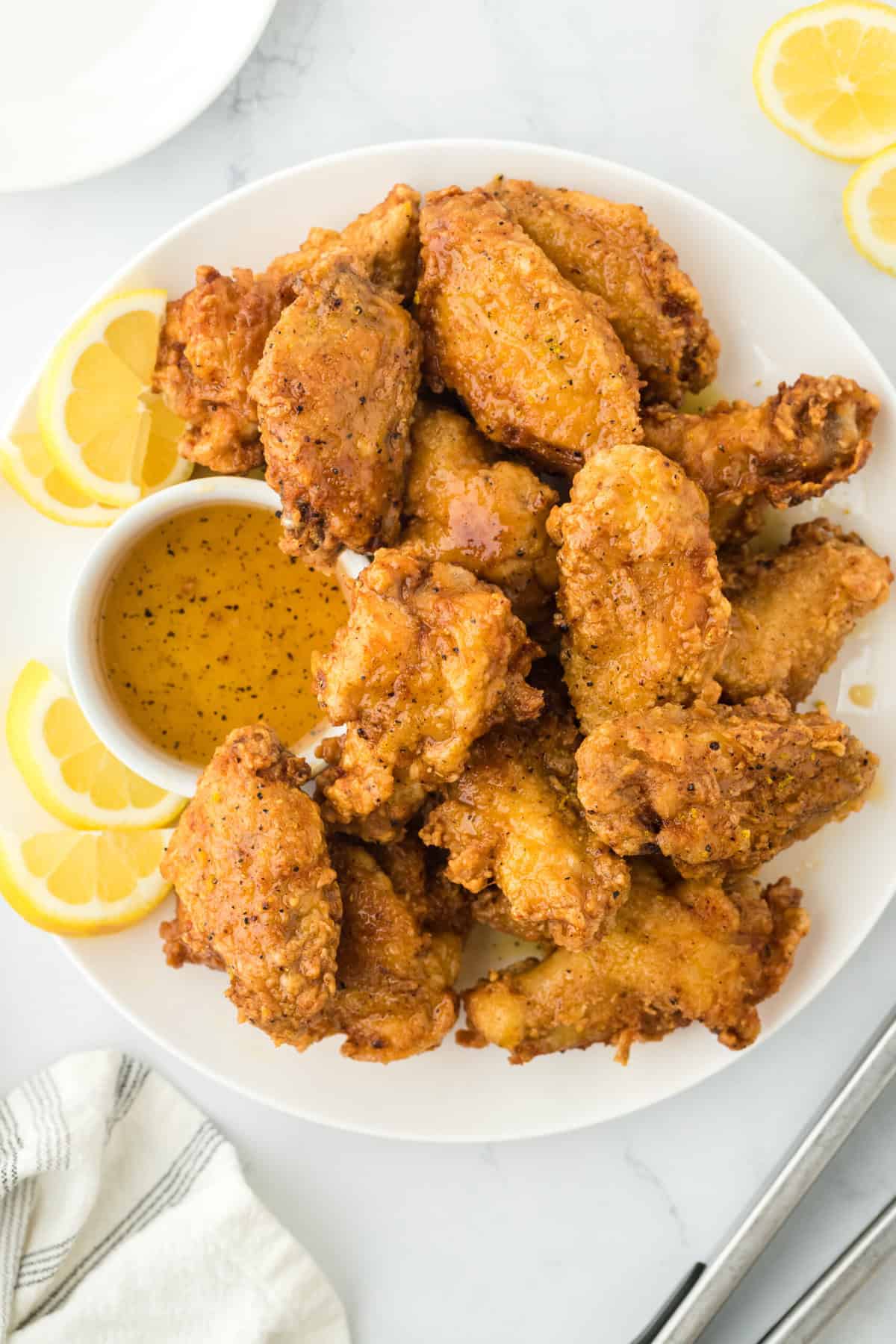 Fried lemon pepper wings on a white plate with lemon wedges and honey lemon pepper wet sauce in a bowl against white background