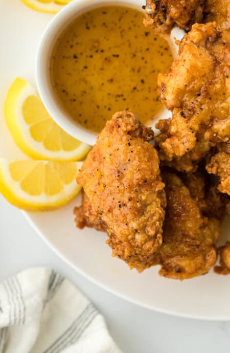 A close up of a lemon pepper chicken wings recipe on a white plate with lemon wedges and sauce on white background