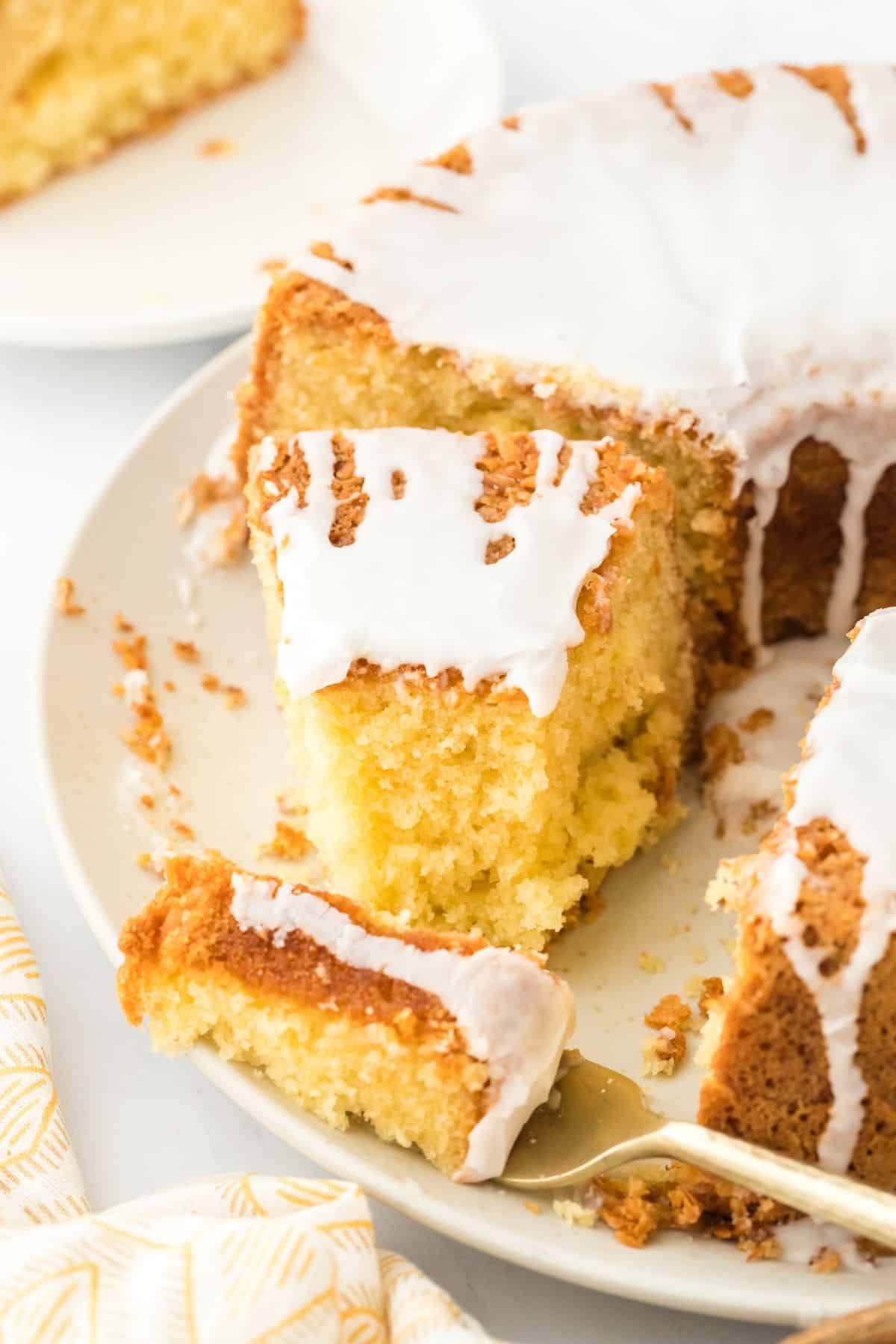 A slice of Louisiana Crunch Cake with glaze on a plate, with a fork lifting a portion, and the rest of the cake next to it