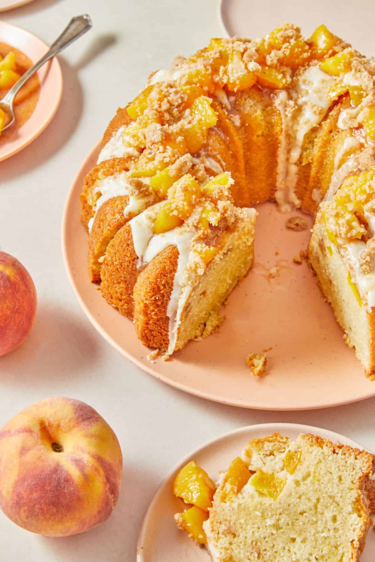 A sliced peach cobbler pound cake with one piece removed, showing the cakeโs interior. The cake is garnished with peach chunks and crumble topping, drizzled with glaze, and accompanied by whole peaches, all set on a pink plate