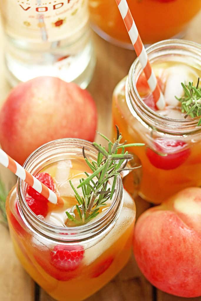 Close up of an overhead shot of two glass mugs filled with Peach Punch garnished with fresh raspberries and striped straws and a pitcher of the punch and a bottle of vodka in the background