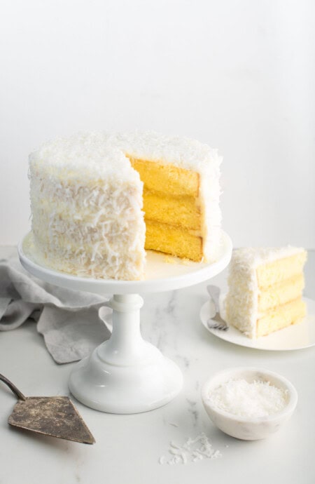 Pineapple Coconut Cake recipe on white cake stand with slice of cake on white plate against white background