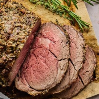 Sliced prime rib on a wooden cutting board with rosemary nearby