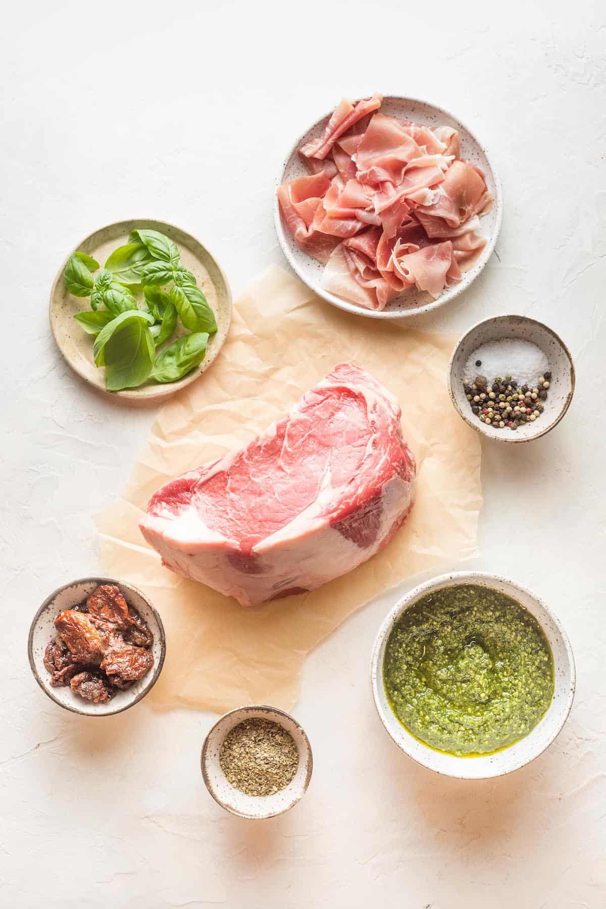 Ingredients to make prosciutto wrapped beef roast in bowls on a white surface.