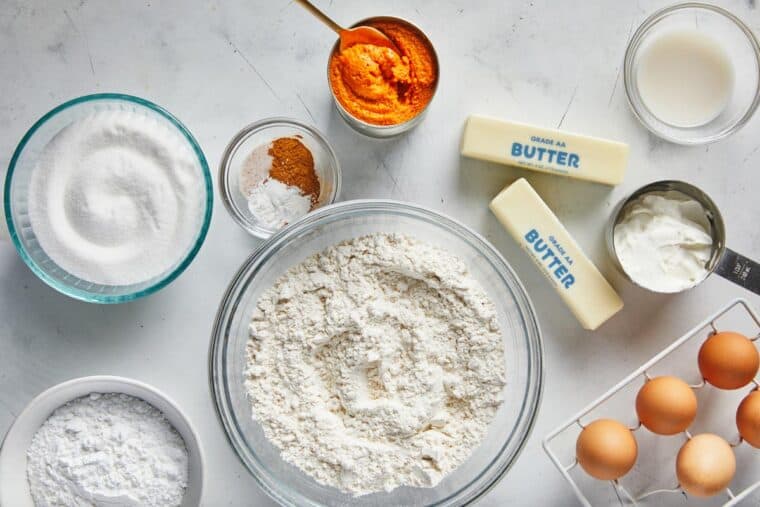 Flour, sugar, pumpkin, spices and eggs to make a bundt cake on a white countertop.