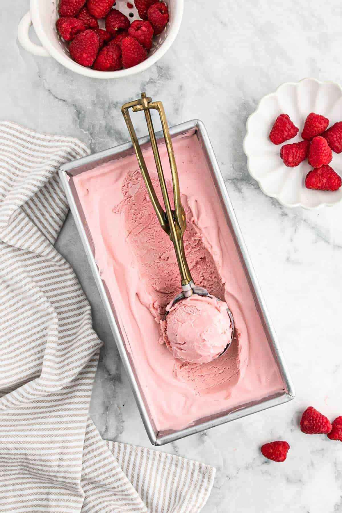 raspberry ice cream Raspberry ice cream in a loaf pan with a scoop inside of an ice cream scoop and bowls of fresh raspberries to the side.