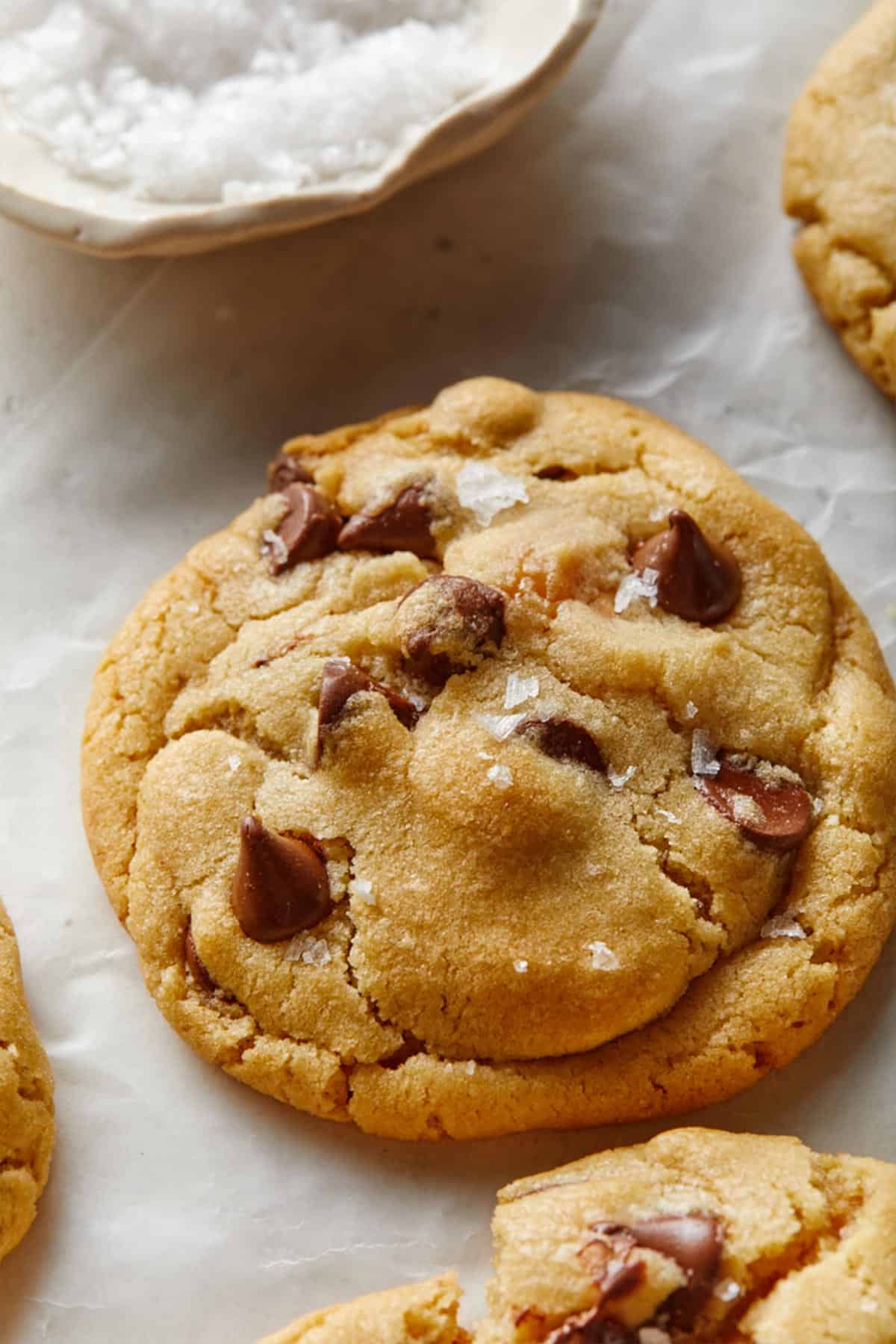 A salted caramel chocolate chip cookie on a baking tray with other cookies sprinkled lightly with flaky sea salt.