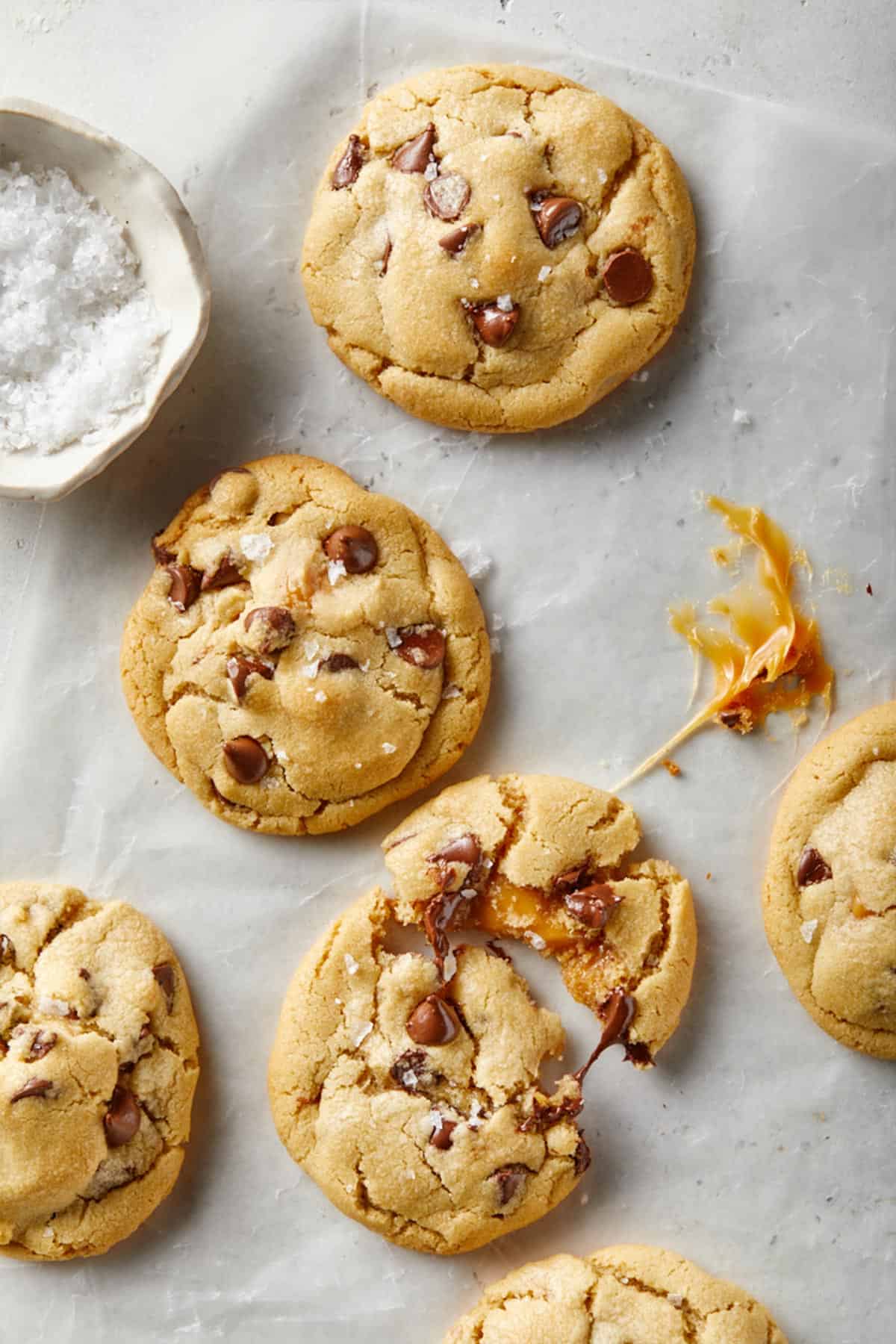 Close up of a salted caramel chocolate chip cookie pulled in half displaying the caramel inside with more cookies on the baking pan.