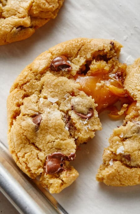 A salted caramel chocolate chip cookie broken in half with caramel oozing out on the baking pan.