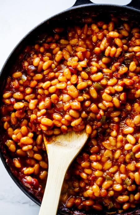 A large wooden spoon serving baked beans in a large skillet