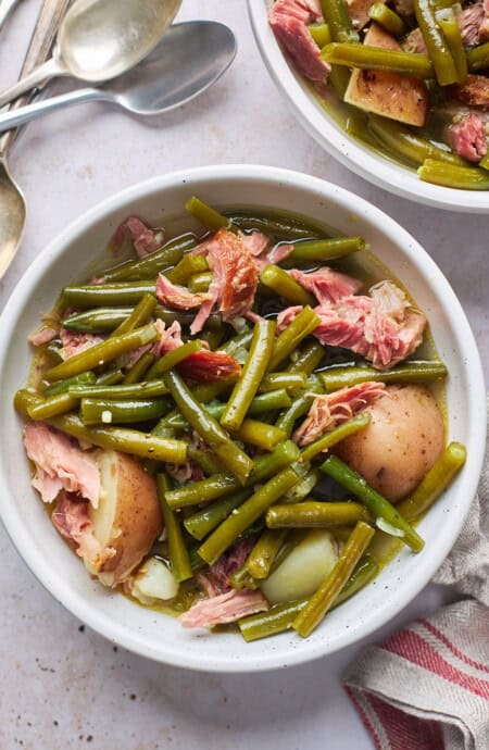 Two bowls of Southern style green beans with potatoes in them with ham hock on a white background