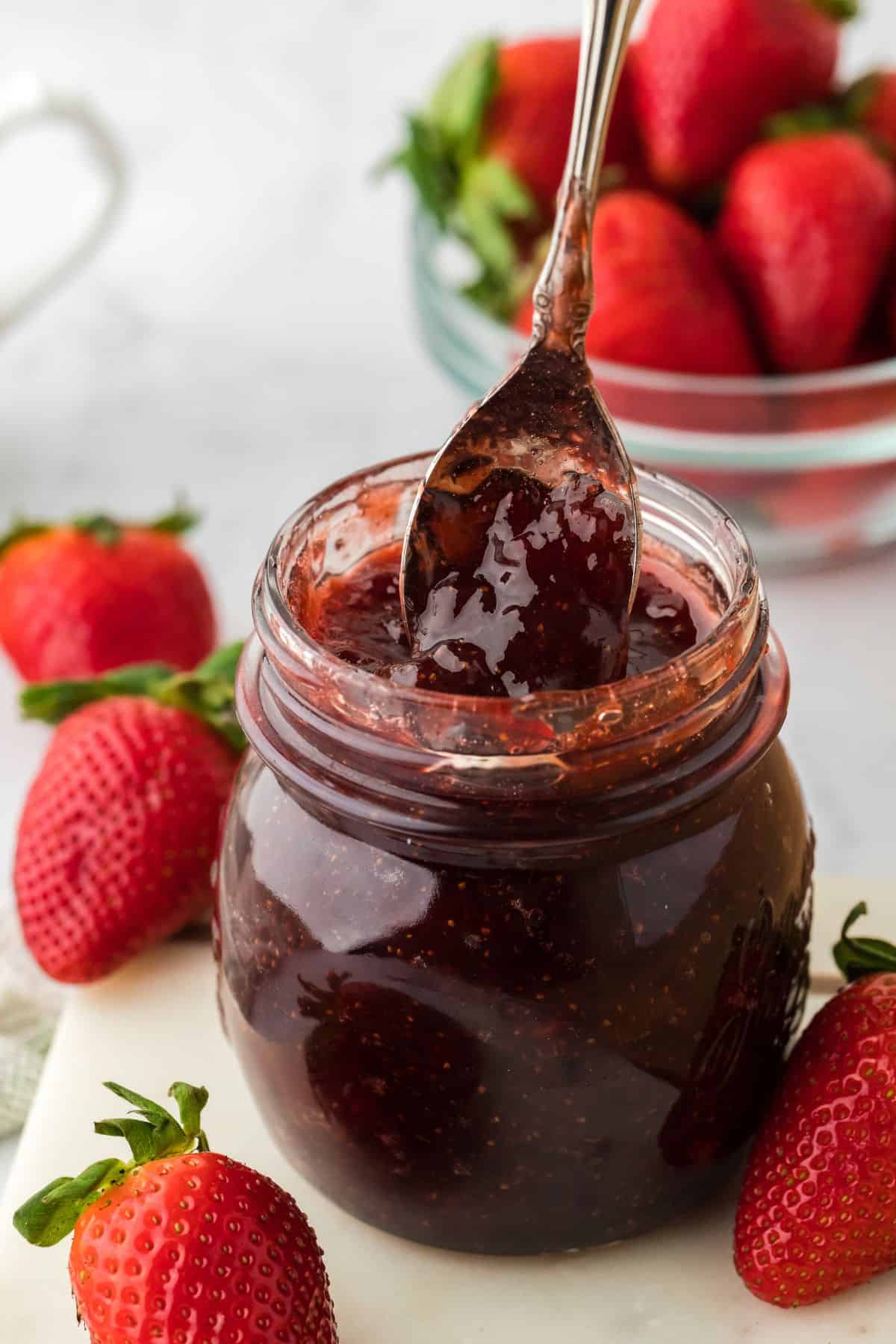 A spoonful of rich strawberry preserves is being lifted from a glass jar, with whole ripe strawberries and a bowl of strawberries in the background