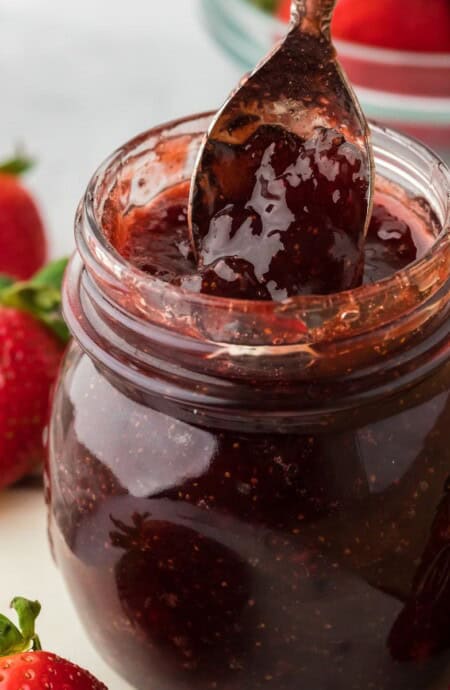 A spoonful of rich strawberry preserves is being lifted from a glass jar, with whole ripe strawberries and a bowl of strawberries in the background