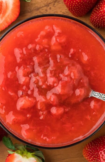 Overhead shot of a bowl of freshly made strawberry sauce with a spoon resting in the bowl. Surrounding the bowl are several strawberries