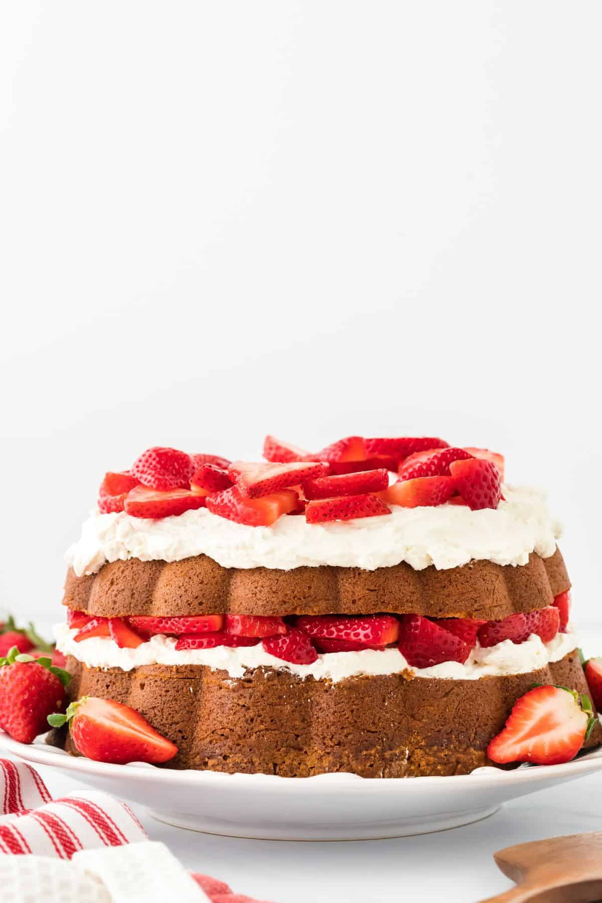 Strawberry shortcake pound cake with fluffy whipped cream and sliced strawberries topping each layer, displayed on a white plate with additional strawberries placed around it