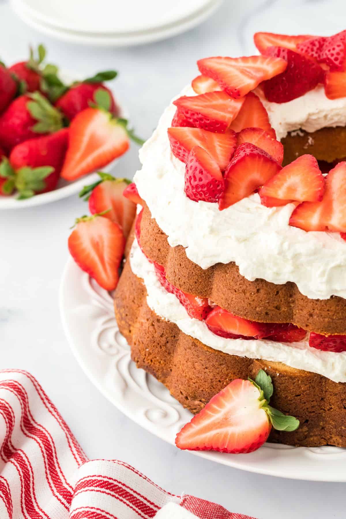 Closeup of strawberry shortcake pound cake showing the layers of golden-brown pound cake with whipped cream and fresh strawberry slices. The cake is placed on a white plate, with more strawberries and a red and white striped towel in the background