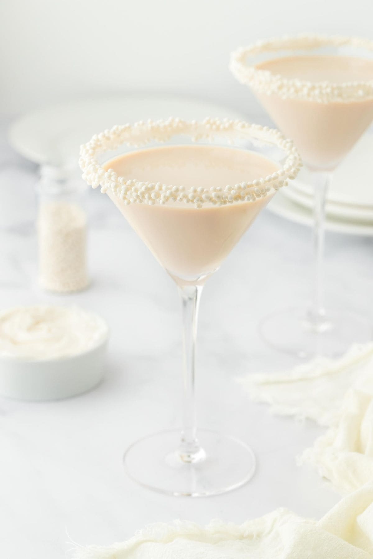 Two martini glasses with sugar pearl-sprinkled rims filled with sugar cookie martini, alongside a small bowl of white frosting