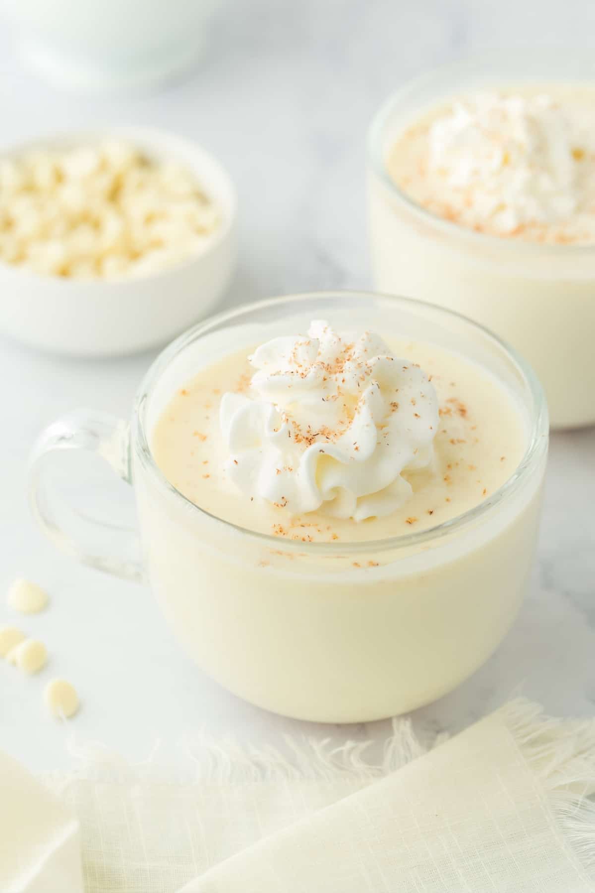 A mug of white hot cocoa topped with whipped cream and some nutmeg on the table.