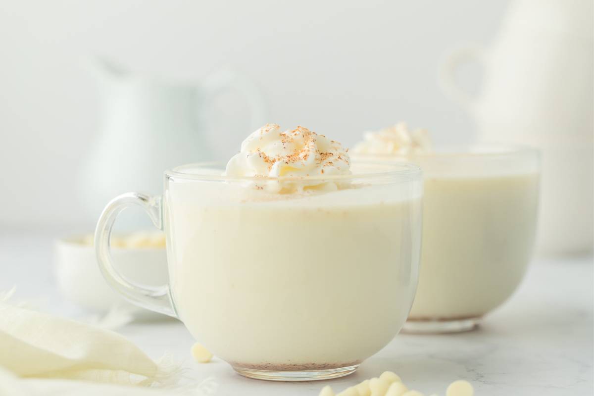A mug of white hot chocolate on the table with a jar of milk in the background.